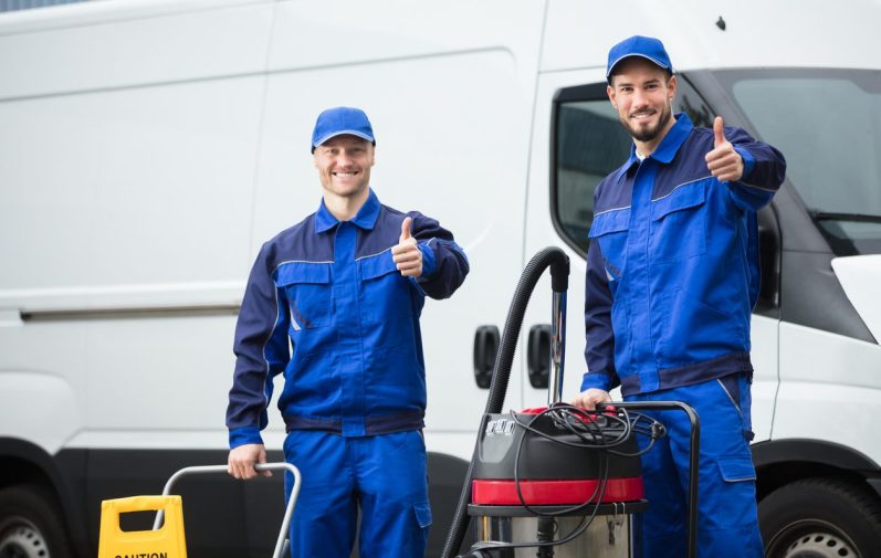 Zwei Männer in blauen Arbeitsanzügen zeigen Daumen nach oben neben einem Transporter.