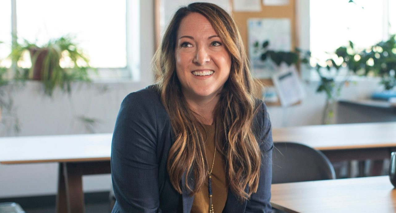 Lächelnde Frau mit langen Haaren in einem hellen, begrünten Raum.